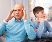 A frustrated man holds his head in his hands, sitting next to a teenage boy who is turned away with his arms crossed.