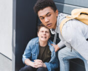 Two teenage boys—one sitting upset on the ground, the other helping him and looking concerned.