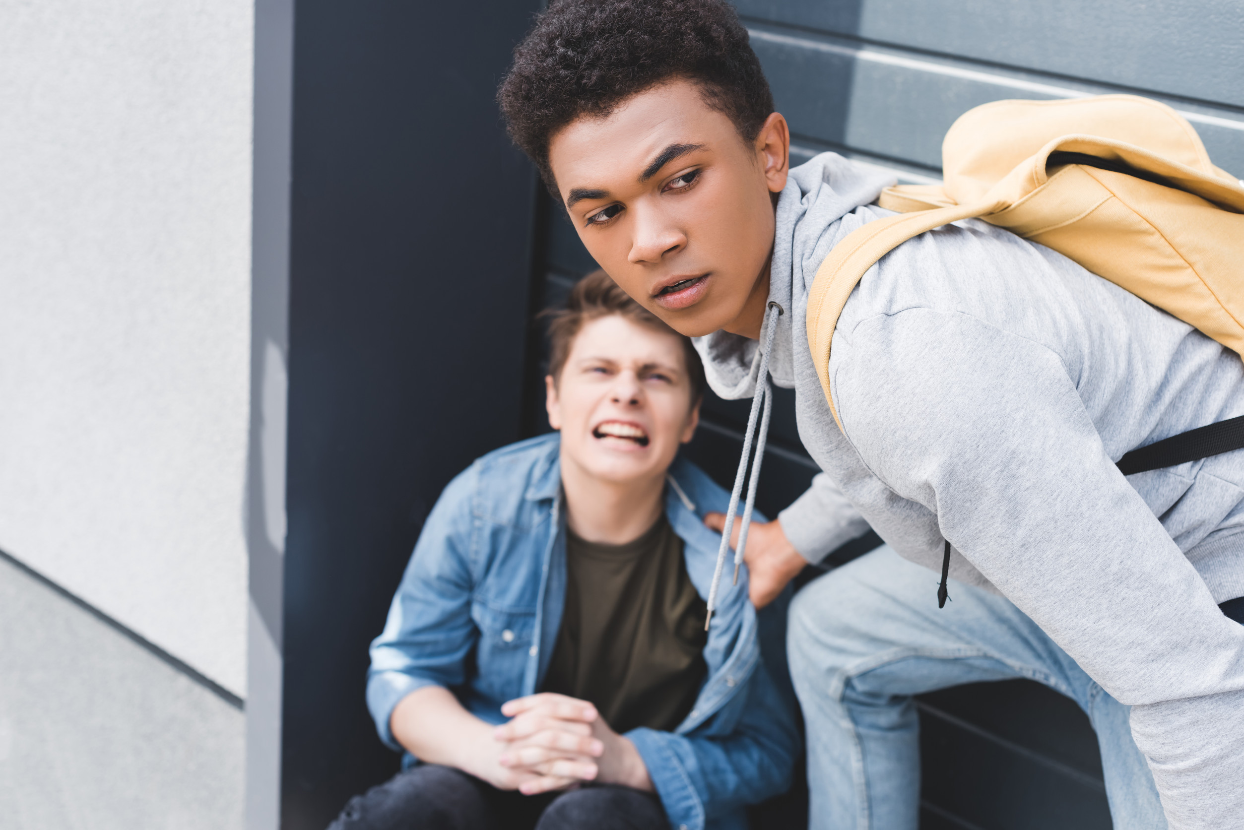 Two teenage boys—one sitting upset on the ground, the other helping him and looking concerned.