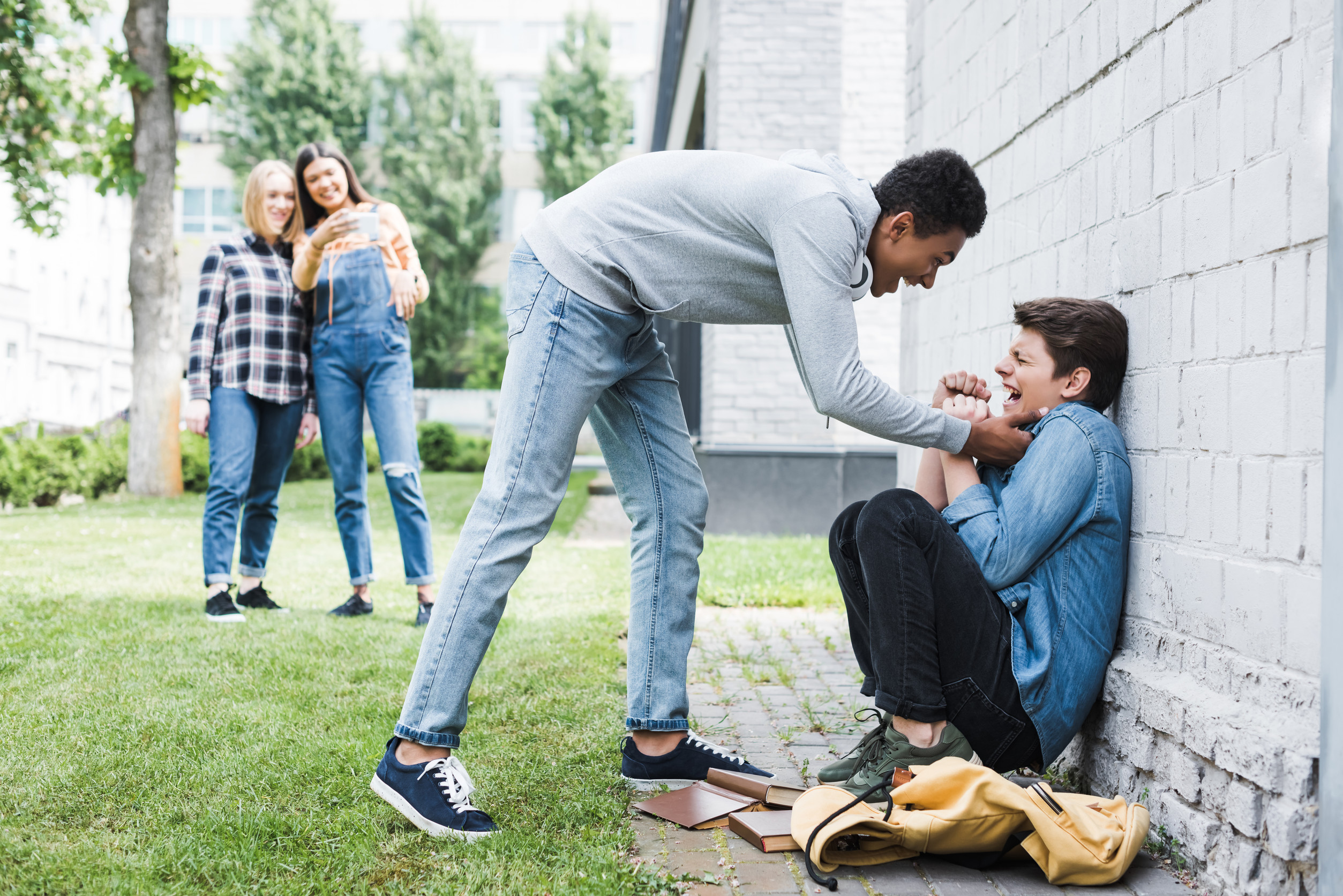 A teenage boy pushes another against a wall while two girls nearby watch and laugh.