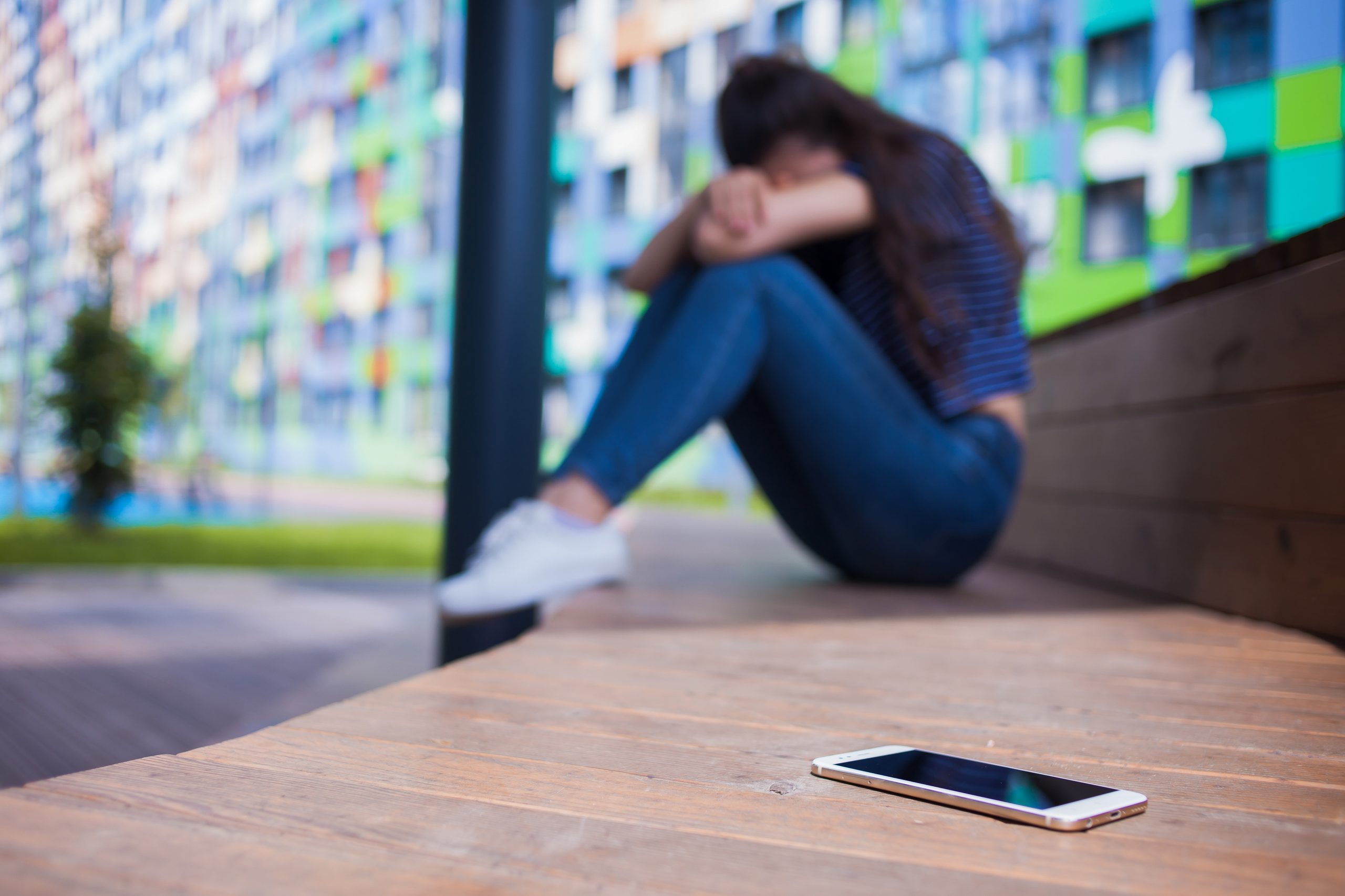 Smartphone lying in the foreground, on blurred background, girl, hiding face in her knees, sitting on a wooden bench.