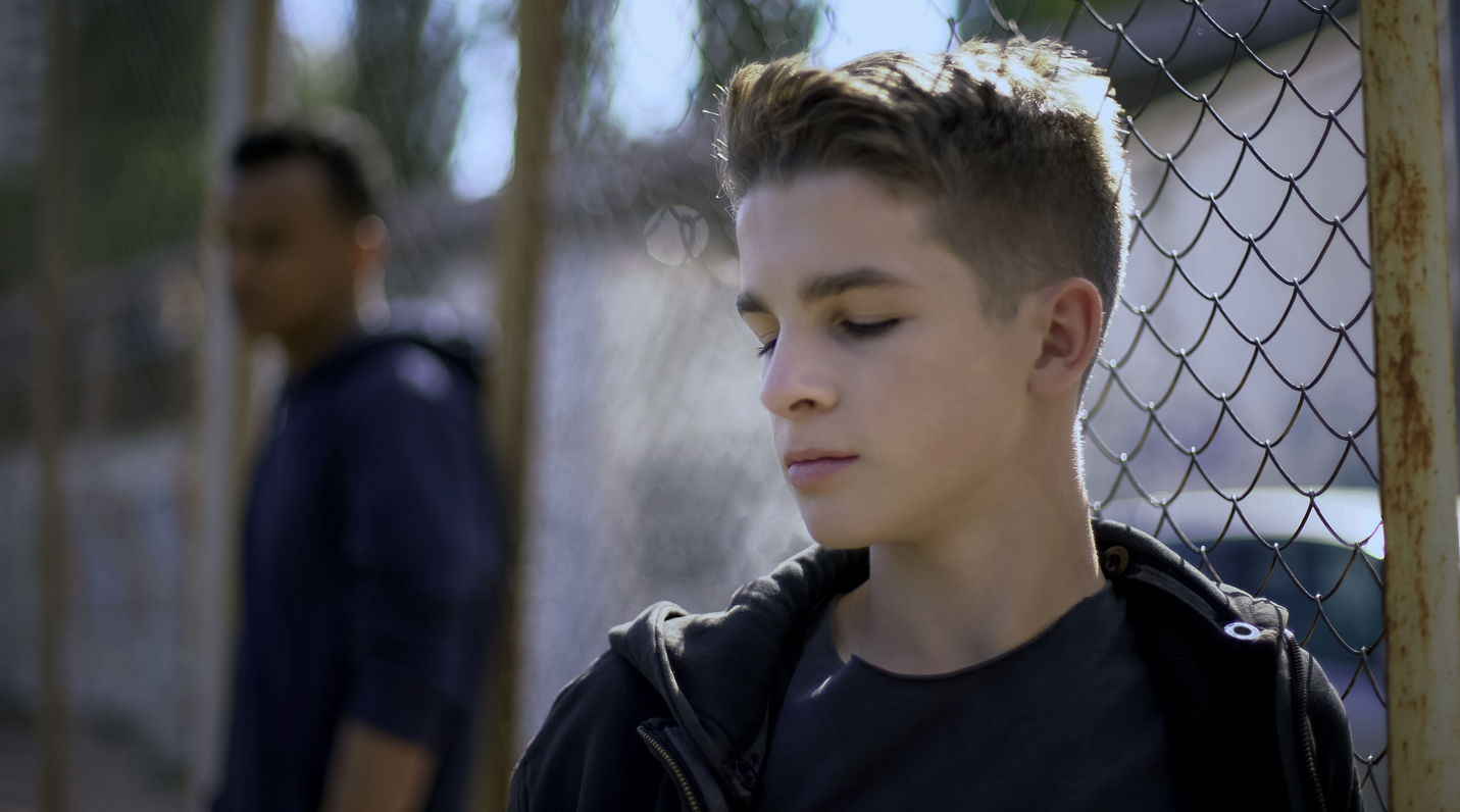 A teenage boy standing in front of a fence looking downcast.