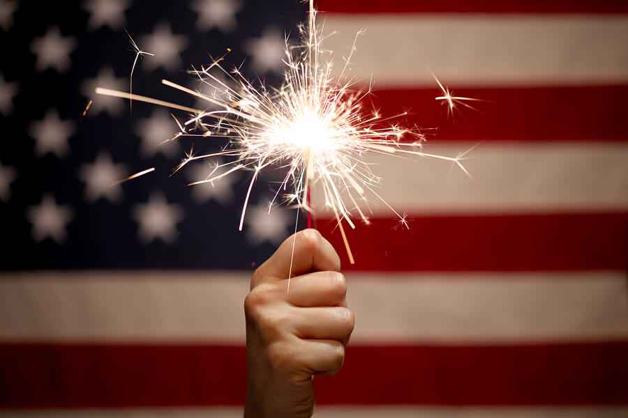 Hand holding lit sparkler in front of the American Flag for 4th